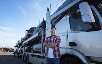Portrait of professional smiling truck driver with crossed arms transporting cars to the market. In background truck trailer with cars.